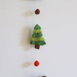 A charming green felt tree, part of a felt forest animal garland, is displayed against a plain white wall. Crafted from soft, textured 100% wool felt, this needle-felted tree features three 3D layers in varying green shades, supported by a brown trunk. A thin white string connects it to a small brown felt ball above and a red felt ball with another forest animal below, creating a whimsical forest scene.