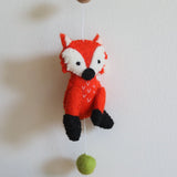 A vibrant orange felt fox, part of a Felt Forest Animal Garland, is suspended against a light, neutral background. This 3D-designed, handmade wool felt fox features white and black accents with small stitched details on its chest, showcasing its textured, needle-felted appearance. A small green felt ball hangs below the fox, completing the cheerful design.