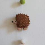 A close-up of a charming wool felt forest animal garland is displayed against a neutral backdrop, featuring a detailed needle-felted hedgehog. The hedgehog is brown with beige accents and tiny dashes mimicking spines. Above, a fuzzy green felt ball connects to the garland, while below, a white felt ball with a copper bell hangs from a thin white string.