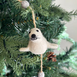 A white seal felt garland is displayed on a festive Christmas tree, evoking holiday cheer. The handmade felt garland features a white felt seal with a black nose and eyes, its flippers extended, strung on a natural fiber string. Two fuzzy grey felt balls accent the string above and below the seal. The garland rests among the green needles of the tree, subtly lit and adorned with pine cones.