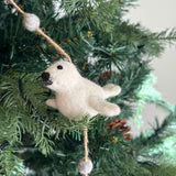 A Seal felt garland is displayed on a green Christmas tree, creating a festive and playful atmosphere. The wool felt garland features three plush, white seals, each with small black eyes and a pronounced dark muzzle. It is strung with natural fiber twine interspersed with gray felt balls, enhancing its soft, cozy appeal.
