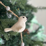 A felt seal garland is draped on a green Christmas tree, setting a cheerful, festive mood. This marine-inspired wool felt garland features multiple charming seals. One prominent light beige felt seal has a small black nose and eyes, adding a whimsical touch. It hangs from a thin, light brown string with small gray felt balls above and below.