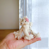 A white poodle wool felt keychain is held in a female hand, set against a softly blurred white background. The handmade keychain features a textured, wool-like poodle with a pink spot on its head, black eyes, and a metal key ring, highlighting its artisanal quality.