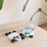 Two wool felt panda coasters rest on a light wooden table, one positioned under a clear glass and the other displayed beside it. These charming, handmade panda felt coasters feature a whimsical black and white design with rounded ears and subtle needle-felted facial details, highlighting their ethical craftsmanship.