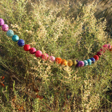 A vibrant wool felt Rainbow Pompom Ball Garland is gracefully draped amidst the light green and tan foliage of a natural shrub. The colorful garland stretches horizontally, featuring soft wool felt balls in shades of purple, pink, red, orange, yellow, green, blue, and turquoise. Its bright, soft texture beautifully contrasts with the shrub's natural, rigid leaves, creating a soft and natural scene.