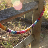 A colorful wool felt rainbow pompom ball garland is draped across a rustic wooden fence on a sunny day. The garland features a series of round felt balls in a vibrant rainbow spectrum of red, orange, yellow, green, blue, indigo, and violet. Each ball is needle-felted, adding texture and a playful 3D design.