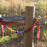 A Rainbow Pompom Ball Wool Felt Garland is gracefully draped over a weathered wooden post outdoors, set against a soft background of tall grass and foliage. The garland features a string of colorful, 3D-designed wool felt balls in vibrant rainbow hues: red, orange, yellow, green, blue, and purple. These soft, slightly fuzzy felt balls are evenly spaced, adding a playful and cheerful touch to the rustic scene.