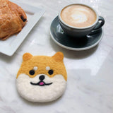 A handmade felt Shiba Inu dog coaster is presented on a marble surface next to a gray coffee cup and a chocolate pastry on a white plate, creating a cozy scene. This handcrafted wool felt Shiba Inu coaster features golden and white wool, with small round ears and a smiling face detailed with black eyes and a pink tongue. Its needle-felted design is ethically handmade by Nepalese female artisans, adding a unique and inviting touch to any home decor.