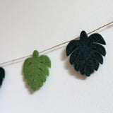 A Greenish Monstera Leaf felt garland, a nature-inspired decor piece, is shown in a close-up, hanging against a stark white wall. The felt garland features evenly spaced monstera leaves on a thin jute string. Each soft leaf alternates between light and dark green, with a textured surface mimicking natural veins.