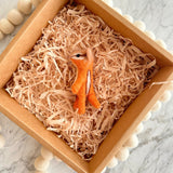 A handcrafted wool felt hamster finger puppet is nestled in a light brown cardboard box, resembling a cozy nest filled with shredded paper. The box, a unique felt toy display, is adorned with a string of small white felt balls along its edges. This orange and cream hamster finger puppet features a white belly stripe, pointed ears, stitched paws, and a black bead eye, ready for imaginative play.