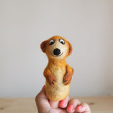 A needle felted meerkat finger puppet is presented in a close-up on a wooden surface. This handmade felt meerkat finger puppet is crafted in light brown and cream hues, featuring diminutive rounded ears, a charming black smile, a pointed snout, and a bushy tail. Its whimsical character is captured in a playful pose.