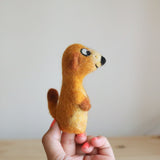 A handmade felt meerkat finger puppet is shown held by a woman's hand against a soft, neutral backdrop. This charming needle felted meerkat puppet, crafted from soft felt, features light-colored fur, a pointed snout, and a bushy tail, capturing the animal's curious and playful stance. The woman's hand, adorned with coral nail polish, highlights the intricate craftsmanship of this unique finger puppet.