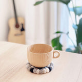 A circular neutral gray felt ball coaster, handcrafted from 100% organic lamb wool, sits on a light wooden surface with a matching light-wood cup on top. This unique handmade felt coaster, composed of hand-stitched gray felt balls, evokes a serene home atmosphere. Ethically made by Nepalese female artisans, it is complemented by a guitar and greenery in the soft background.