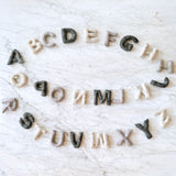 A vibrant wool felt Rainbow Alphabet Banner Kids Room Garland is displayed on a white marble surface. This garland features all 26 letters of the alphabet, each crafted in bright, eye-catching rainbow colors and strung together by a thin white string.