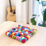 A vibrant Rainbow Square Felt Ball Pot Trivet rests on a light wooden surface, creating a warm, playful atmosphere. This handmade organic wool felt trivet features carefully stitched felt balls in shades of red, orange, yellow, green, blue, purple, pink, brown, and white, colored with azo-free dye. A soft-focus guitar and plants in the background suggest a cozy, creative home setting.