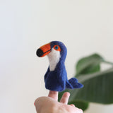 A handmade needle-felted wool toucan finger puppet is playfully displayed on a model's finger against a bright, airy background with a hint of greenery. This unique toucan puppet features a rich dark blue body, a white throat, an orange and black beak, and small blue eyes, showcasing its detailed 3D design.