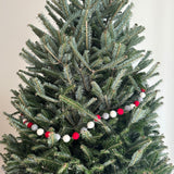 A festive felt pompom ball garland, in alternating red, gray, and white, is draped across the branches of a full green Christmas tree. This felt garland creates a cozy and cheerful atmosphere, with its bright colors contrasting against the tree's textured needles.