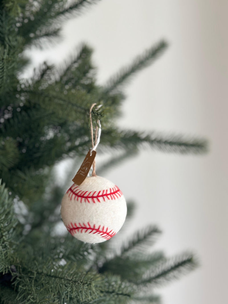 A needle-felted baseball ornament hangs from a lush green Christmas tree. This cheerful, 3-inch wool felt ornament is white with red stitches, replicating a real baseball design. It is suspended by a simple jute string, perfect for holiday decorations or for sports enthusiasts.