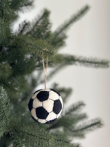 A spherical wool felt soccer ball ornament from the American Popular Sports Ornament Set hangs on a blurred Christmas tree. It features alternating black and white pentagons, crafted from soft, textured 100% wool felt. A jute string is attached for graceful hanging.