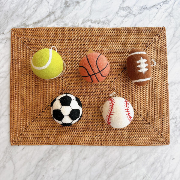 A set of five wool felt American popular sports ball ornaments rests on a woven mat against a marble background. These 3-inch, 3D needle felted ornaments include a yellow tennis ball, orange basketball, brown football with white accents, classic black and white soccer ball, and white baseball with red stitching. Each handcrafted sports ornament has a jute thread for easy hanging.