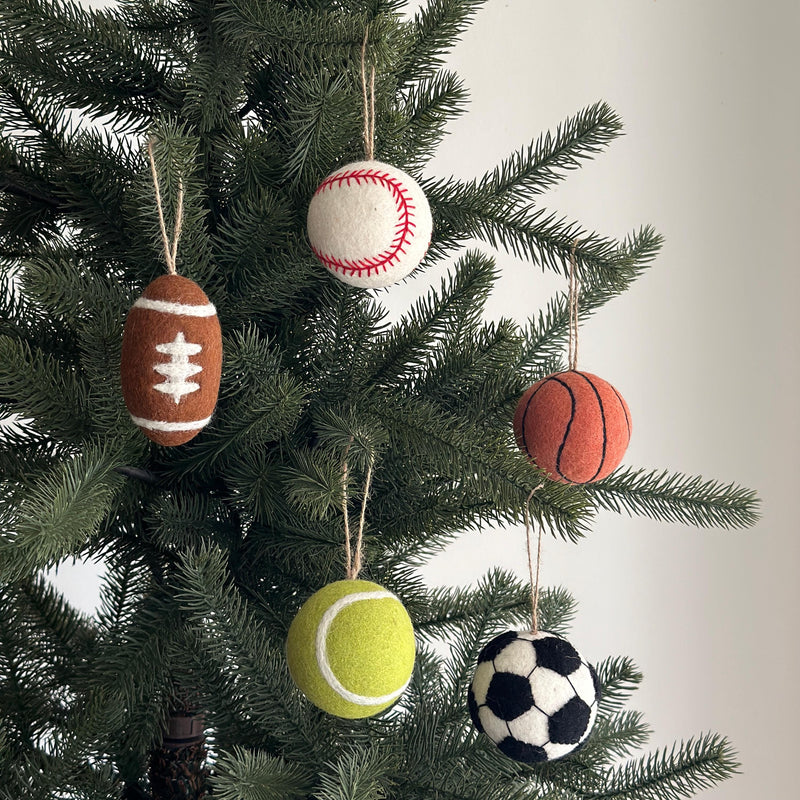 A set of five handcrafted wool felt American sports ball ornaments hang from lush green Christmas tree branches. Each unique ornament, suspended by a jute string, includes a brown American football with white accents, a white baseball with red stitching, an orange basketball with black lines, a bright yellow tennis ball, and a black-and-white soccer ball, adding a festive and sporty touch.