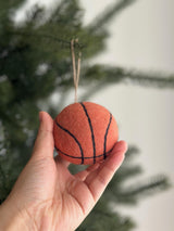 A handmade, needle-felted basketball ornament is gently held against a blurred Christmas tree background. This round, 3-inch orange wool felt ornament features distinct black lines, mimicking a real basketball. A jute string allows it to hang as a unique Christmas tree ornament.