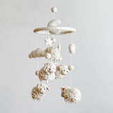 A baby mobile hangs on a wall, featuring a collection of white and cream-colored felt sheep in various sizes. Some sheep have faces, while others are plain. The mobile is suspended from a string, creating a peaceful atmosphere for a nursery.