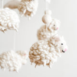 A collection of white, fluffy sheep-shaped objects hang from a string in a semi-circle arrangement, suspended in mid-air. The soft wool felt material and gentle movement create a soothing atmosphere, perfect for a baby's room.