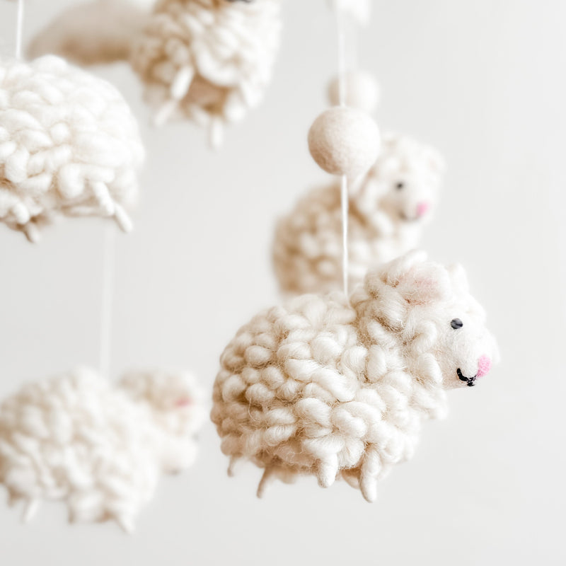 A collection of white, fluffy sheep-shaped objects hang from a string in a semi-circle arrangement, suspended in mid-air. The soft wool felt material and gentle movement create a soothing atmosphere, perfect for a baby's room.