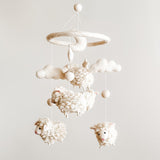 A baby mobile featuring soft, organic wool felt sheep that float gently above, suspended from a circular hoop. The sheep are white with black facial features, creating a serene atmosphere.