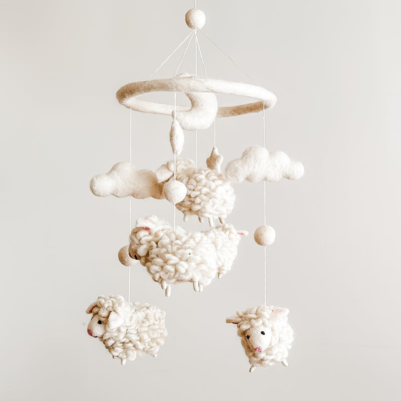 A baby mobile featuring soft, organic wool felt sheep that float gently above, suspended from a circular hoop. The sheep are white with black facial features, creating a serene atmosphere.