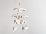 A handcrafted mobile, Baby Mobile - Counting Sheep, hangs from a string against a white background. The mobile features a circular structure at the top with a small figure resembling a sheep or lamb. Below, multiple fluffy, white sheep made of soft wool felt are suspended in a row, each slightly varying in size and position for added visual interest.