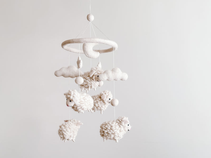 A handcrafted mobile, Baby Mobile - Counting Sheep, hangs from a string against a white background. The mobile features a circular structure at the top with a small figure resembling a sheep or lamb. Below, multiple fluffy, white sheep made of soft wool felt are suspended in a row, each slightly varying in size and position for added visual interest.