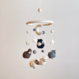A baby mobile, 'Counting Sheep', hangs from a string, featuring a circular white base and a small felt hoop at the top. The mobile includes three sheep figures in different shades of brown, one appearing to move, along with smaller elements resembling stars or moons in various colors.