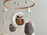 A baby mobile with an owl theme hangs from the ceiling, suspended by strings. The central figure is a wool felt owl, surrounded by smaller felt balls in shades of brown and white. This charming Wool Felt Owl Baby Mobile features a wooden frame and cozy cotton thread, promising endless fun for your little one.