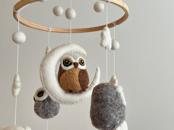 A baby mobile with an owl theme hangs from the ceiling, suspended by strings. The central figure is a wool felt owl, surrounded by smaller felt balls in shades of brown and white. This charming Wool Felt Owl Baby Mobile features a wooden frame and cozy cotton thread, promising endless fun for your little one.