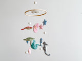 A wooden baby mobile, featuring sea creatures and stars in various colors, hangs from a string against a neutral background. The product is suspended in a real-life setting, ideal for use as a decorative piece in a nursery or child's room to create a peaceful environment.