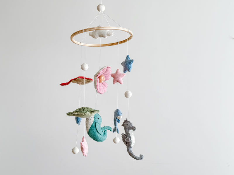 A wooden baby mobile, featuring sea creatures and stars in various colors, hangs from a string against a neutral background. The product is suspended in a real-life setting, ideal for use as a decorative piece in a nursery or child's room to create a peaceful environment.