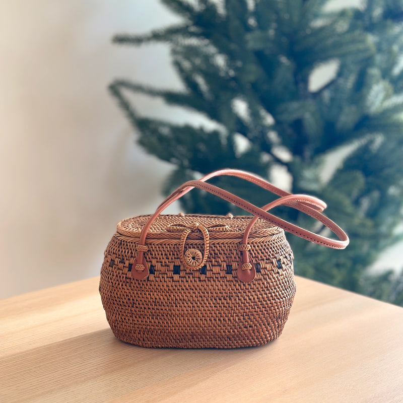 A woven rattan handbag, the Julia, rests on a wooden surface. The oval-shaped bag features a smooth, polished finish with a snag-free design. A genuine leather handle is secured by a bowtie clasp with secure snaps for closure. Lined with Batik cotton, the bag exudes a festive and cozy aesthetic.