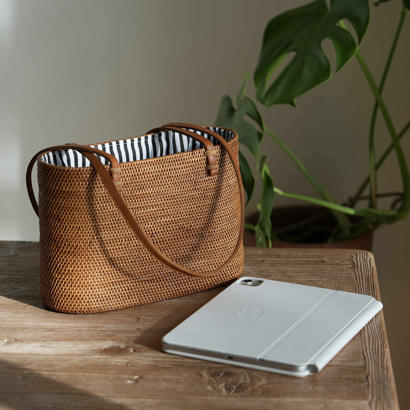 A detailed, eye-level shot of a rectangular, handwoven rattan tote bag resting on a rustic wooden table. The bag, golden-brown in the natural sunlight, features thin, tan leather shoulder straps looped over the sides. The open top reveals a crisp, black-and-white vertically striped fabric lining. In the foreground to the right, a silver tablet with a folded white magnetic cover lies on the table. The scene is illuminated by soft light from the left, casting a gentle shadow on the plain wall behind.