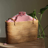 A handwoven rattan tote bag sits in a pool of natural sunlight on a rustic wooden table. The bag features elegant tan leather handles and a vibrant red-and-white striped fabric lining, which is cinched closed with a drawstring at the top. In the background, the soft green leaves of a monstera plant add a touch of nature, complementing the bag's organic materials and exquisite craftsmanship.