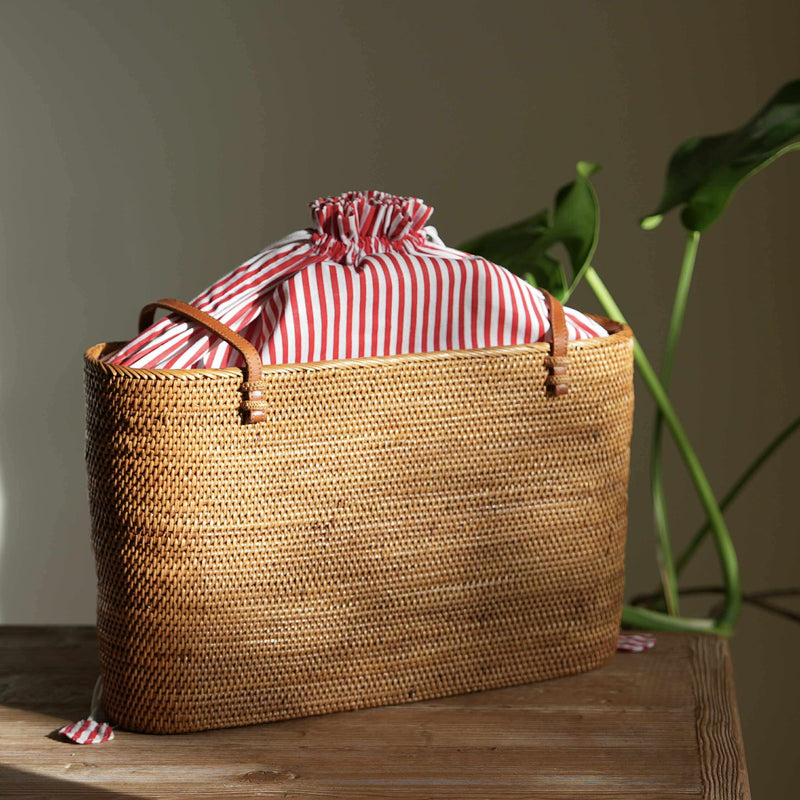 A handwoven rattan tote bag sits in a pool of natural sunlight on a rustic wooden table. The bag features elegant tan leather handles and a vibrant red-and-white striped fabric lining, which is cinched closed with a drawstring at the top. In the background, the soft green leaves of a monstera plant add a touch of nature, complementing the bag's organic materials and exquisite craftsmanship.