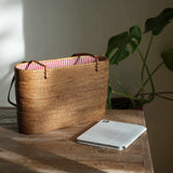 The handcrafted Bali Rattan Shoulder Bag sits elegantly on a rustic wooden table, bathed in natural sunlight. Its sturdy, rectangular shape and intricate weave highlight its artisanal quality, while the vibrant red-and-white striped interior lining adds a playful contrast. Pictured next to a tablet, the spacious tote proves both stylish and functional, perfect for carrying daily essentials. In the background, a monstera plant enhances the scene's natural, relaxed aesthetic.