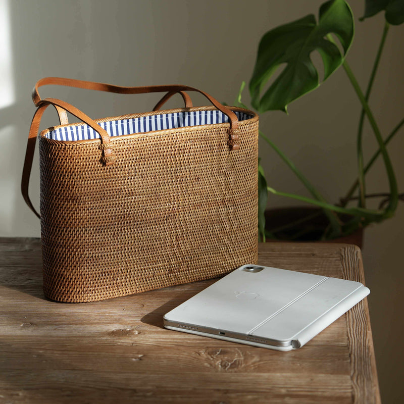 A detailed, eye-level shot of a rectangular, handwoven rattan tote bag resting on a rustic wooden table. The bag, golden-brown in the natural sunlight, features thin, tan leather shoulder straps attached to its rim. The open top reveals a crisp, blue and white vertically striped fabric lining. In the foreground, a silver tablet with a folded white cover lies on the table. To the right, the blurred, deep green leaves of a monstera plant create a natural backdrop, while soft light from the left casts a gentle