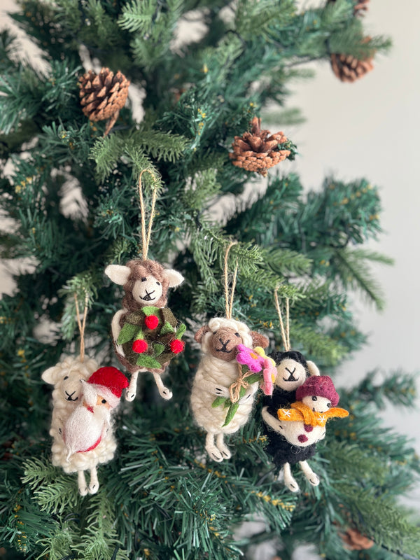 A whimsical black sheep holding snowman felt ornament hangs from a green Christmas tree adorned with natural pine cones. The black sheep ornament is suspended by a jute string and holds a small snowman wearing a red hat and an orange scarf. This handcrafted felt ornament adds a joyful touch to holiday decor.