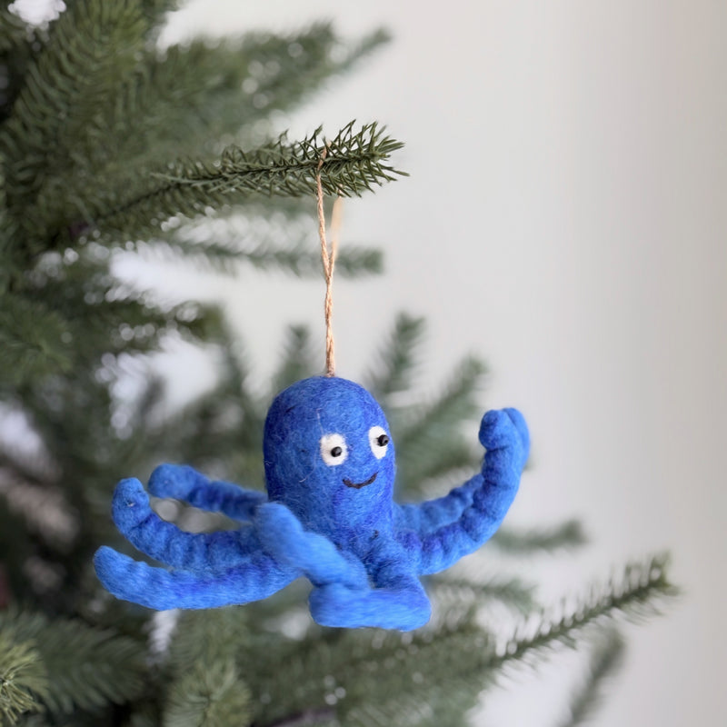 A blue wool felt octopus ornament hangs from a vibrant green pine branch, set against a soft, neutral background. This playful felt octopus features curly tentacles and a cheerful expression with small, round eyes and a stitched smile. A rustic jute string suspends the unique ornament, adding a whimsical touch to the festive scene.