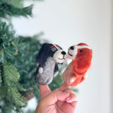 A hand holds two felt finger puppets against a festive Christmas tree background, featuring Cavalier King Charles Spaniel designs. The puppets have detailed features like ears, eyes, noses, and textures mimicking real dogs. They are presented in a playful manner, with the puppets interacting with each other amidst the warm and festive atmosphere.