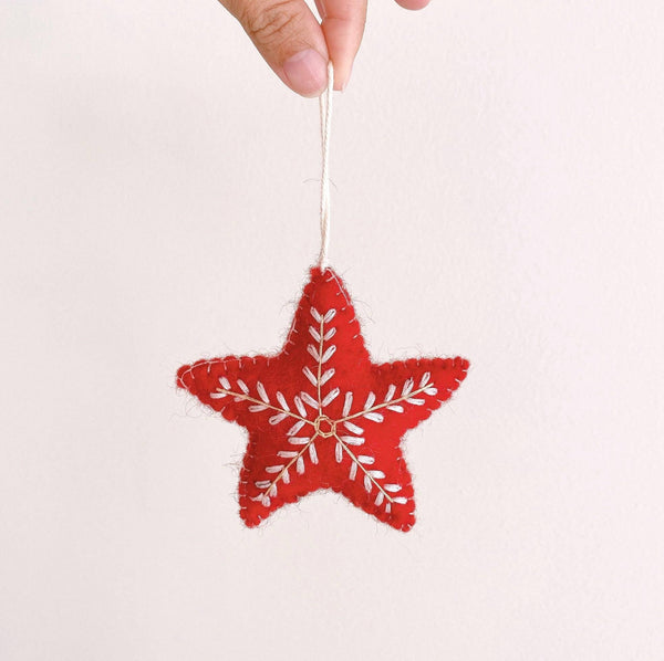 A vibrant red star Christmas ornament is delicately held by a female hand against a plain white backdrop. This handcrafted wool felt star ornament features meticulous white snowflake embroidery and hangs from a white cotton string, evoking winter elegance and holiday charm.