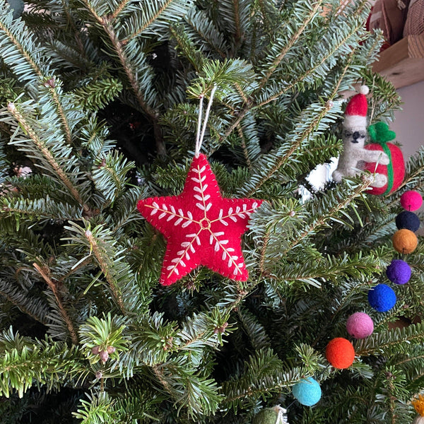 A red wool felt star Christmas ornament hangs on an evergreen Christmas tree. This plump star features delicate white snowflake embroidery on its lightly textured surface and is suspended by a white cotton string.