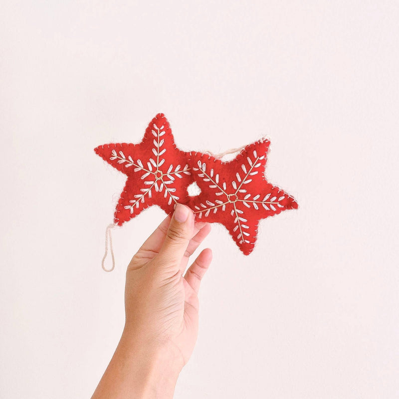 A red wool felt star Christmas ornament is delicately held by a female hand against a soft white backdrop. This handmade ornament features intricate white snowflake embroidery and a simple cotton string for hanging, adding a touch of winter elegance to holiday decor.